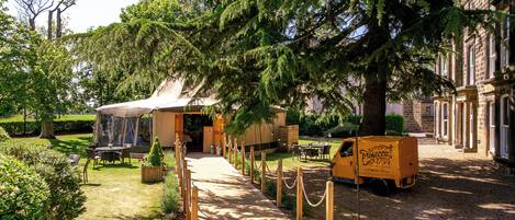 Un patio con una zona cubierta por una carpa, mesas y sillas de madera, y un camión de reparto amarillo.