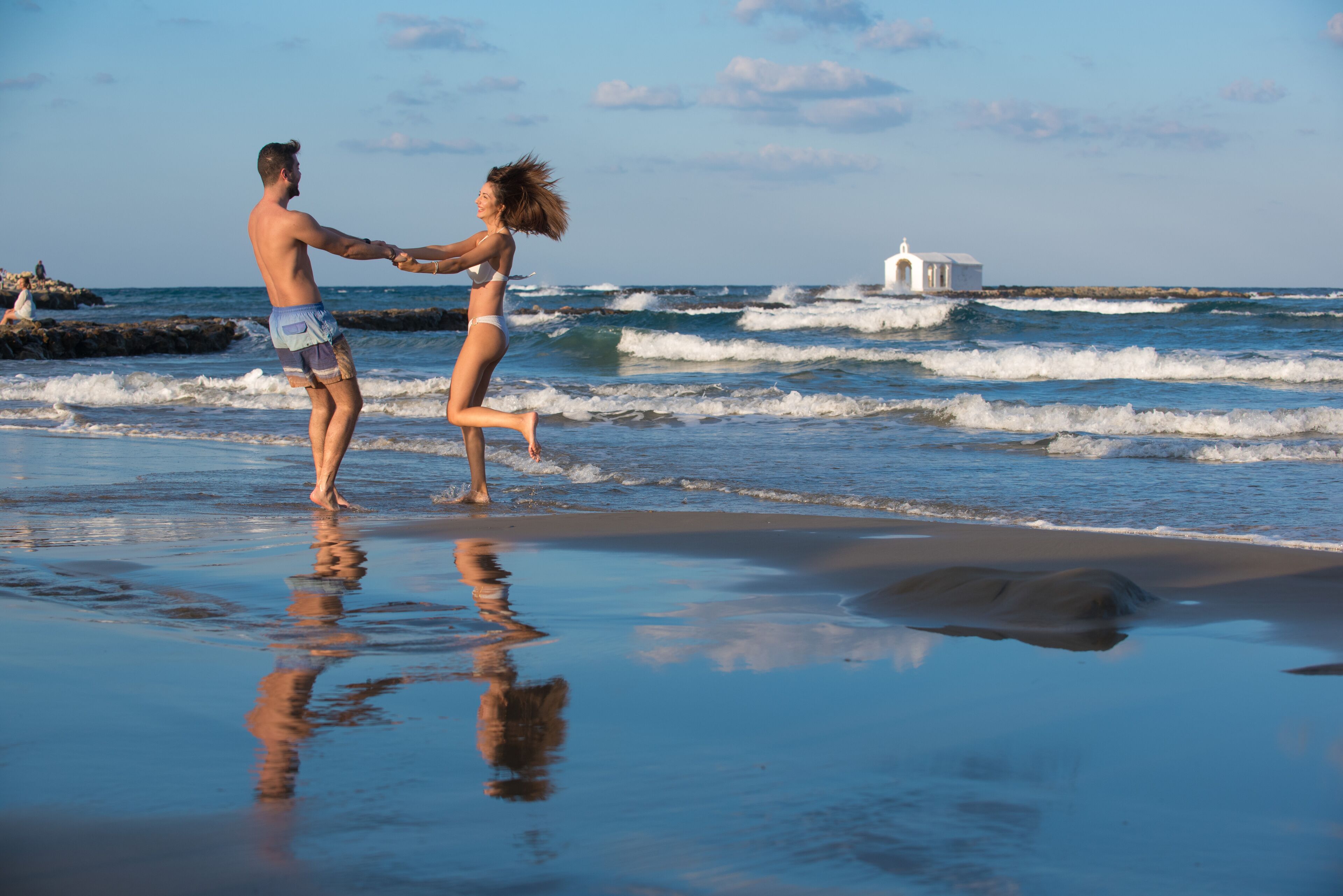 Un couple se tenant la main sur une plage, avec des vagues et un bâtiment blanc en arrière-plan.