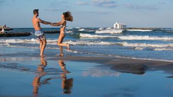 Un couple se tenant la main sur une plage, avec des vagues et un bâtiment blanc en arrière-plan.