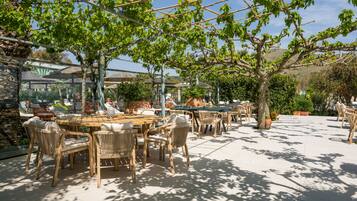 An outdoor patio with round wooden tables and wicker chairs, surrounded by potted plants and a pergola structure.