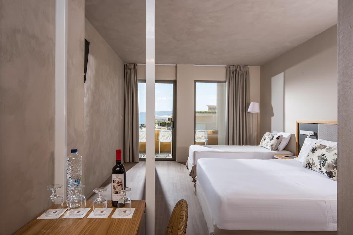 A hotel room with two beds, a wooden table with a bottle of wine and glasses, and a view of the outside balcony.