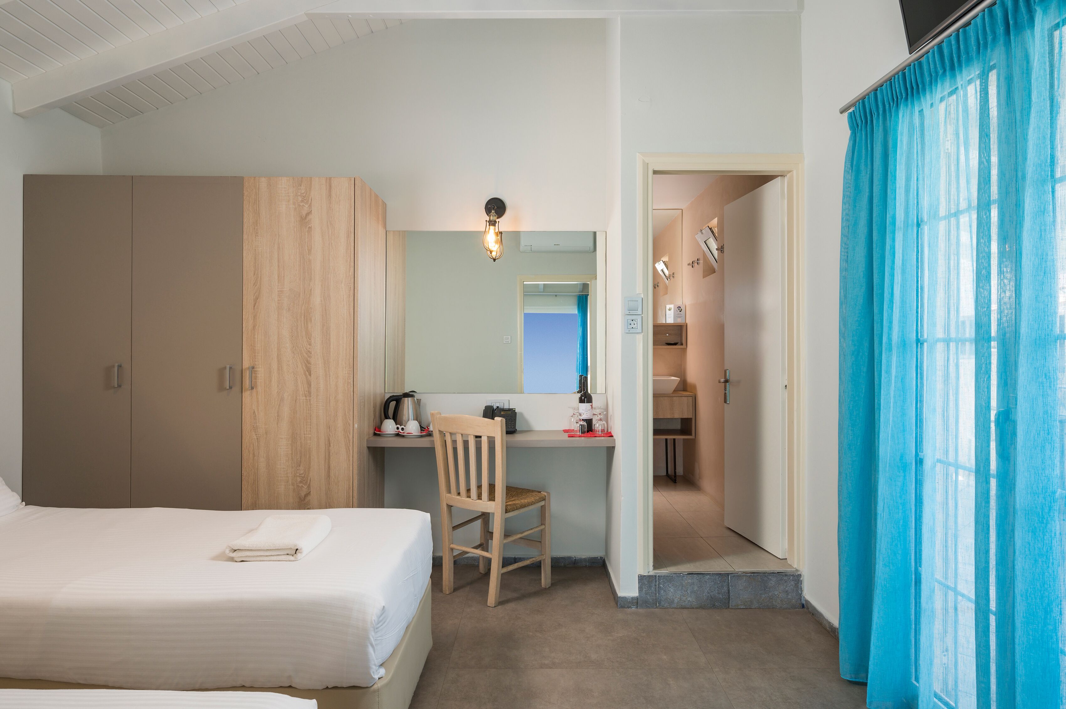 Una habitación de hotel con cama, escritorio con silla, espejo y ventana con cortinas azules.