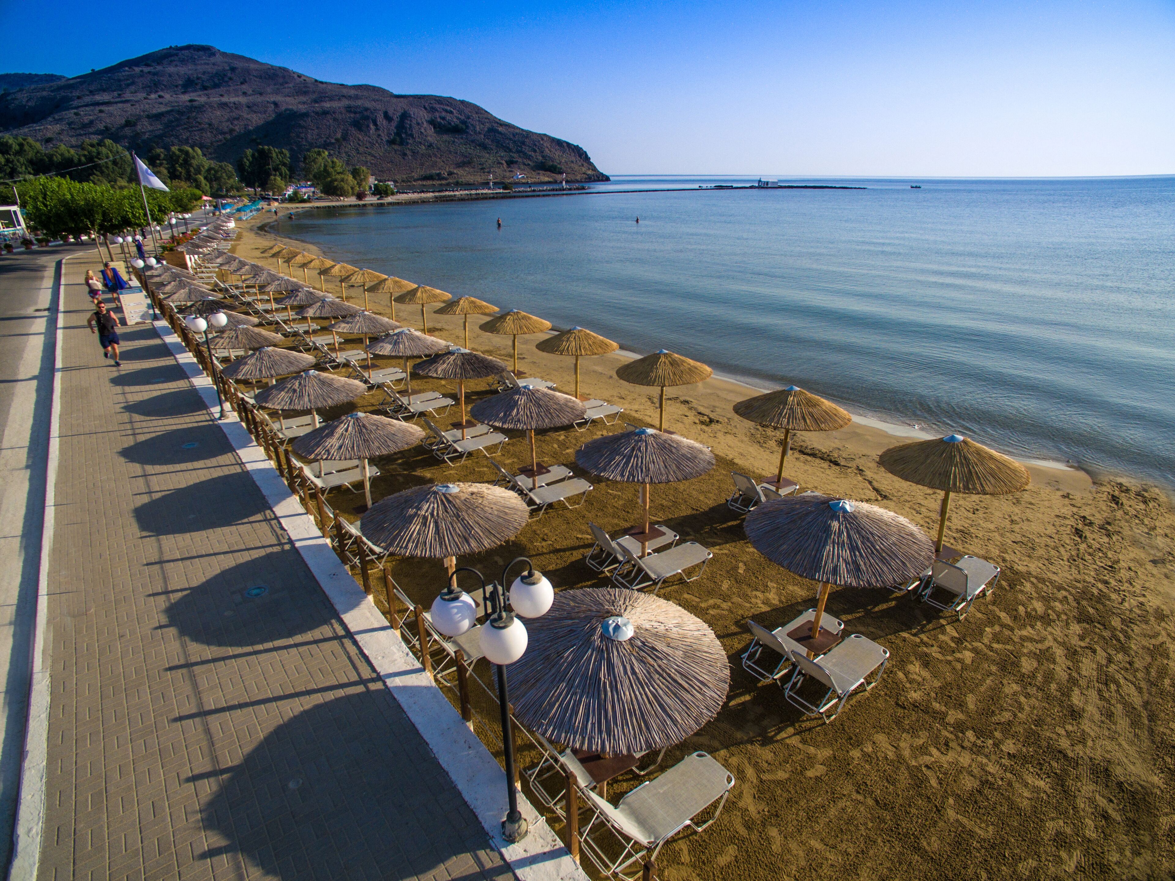 A beach with rows of sun loungers and thatched umbrellas, a paved walkway, and a mountain in the background.