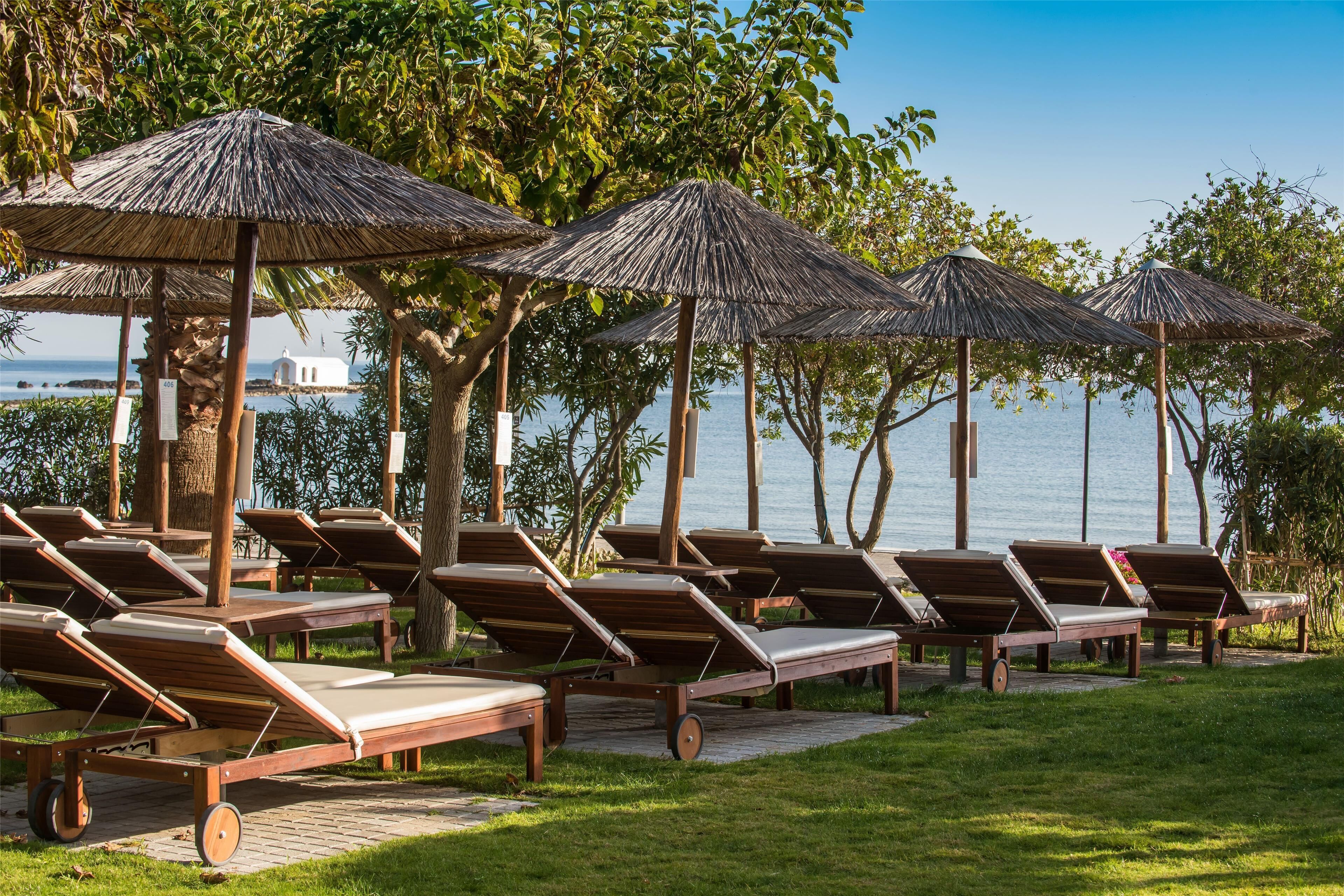 A row of wooden lounge chairs with thatched umbrellas, arranged on a paved area with grass, overlooking a body of water.