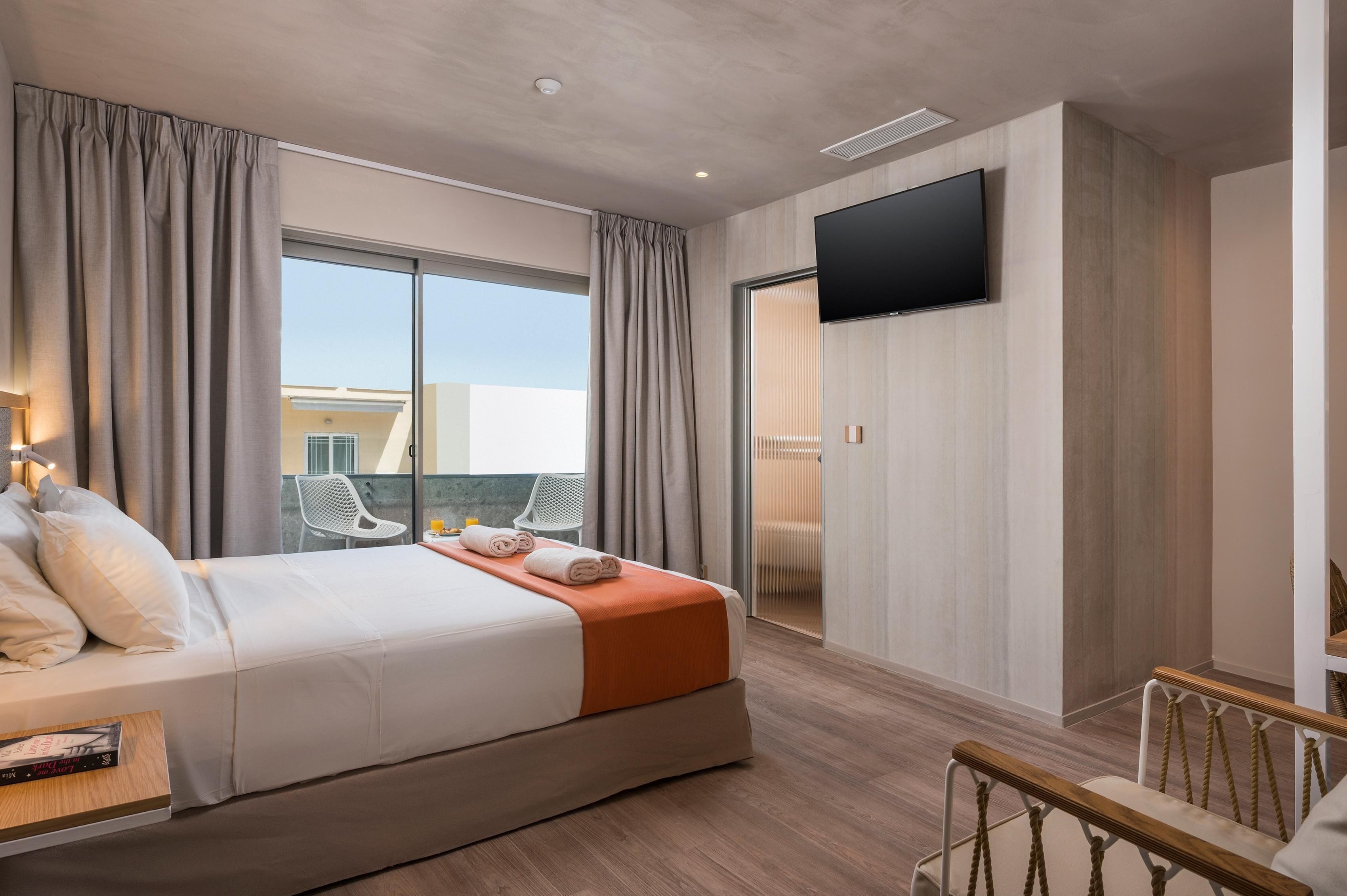 A modern hotel room with a large bed, a flat-screen TV, a balcony with a chair, and a view of the city.