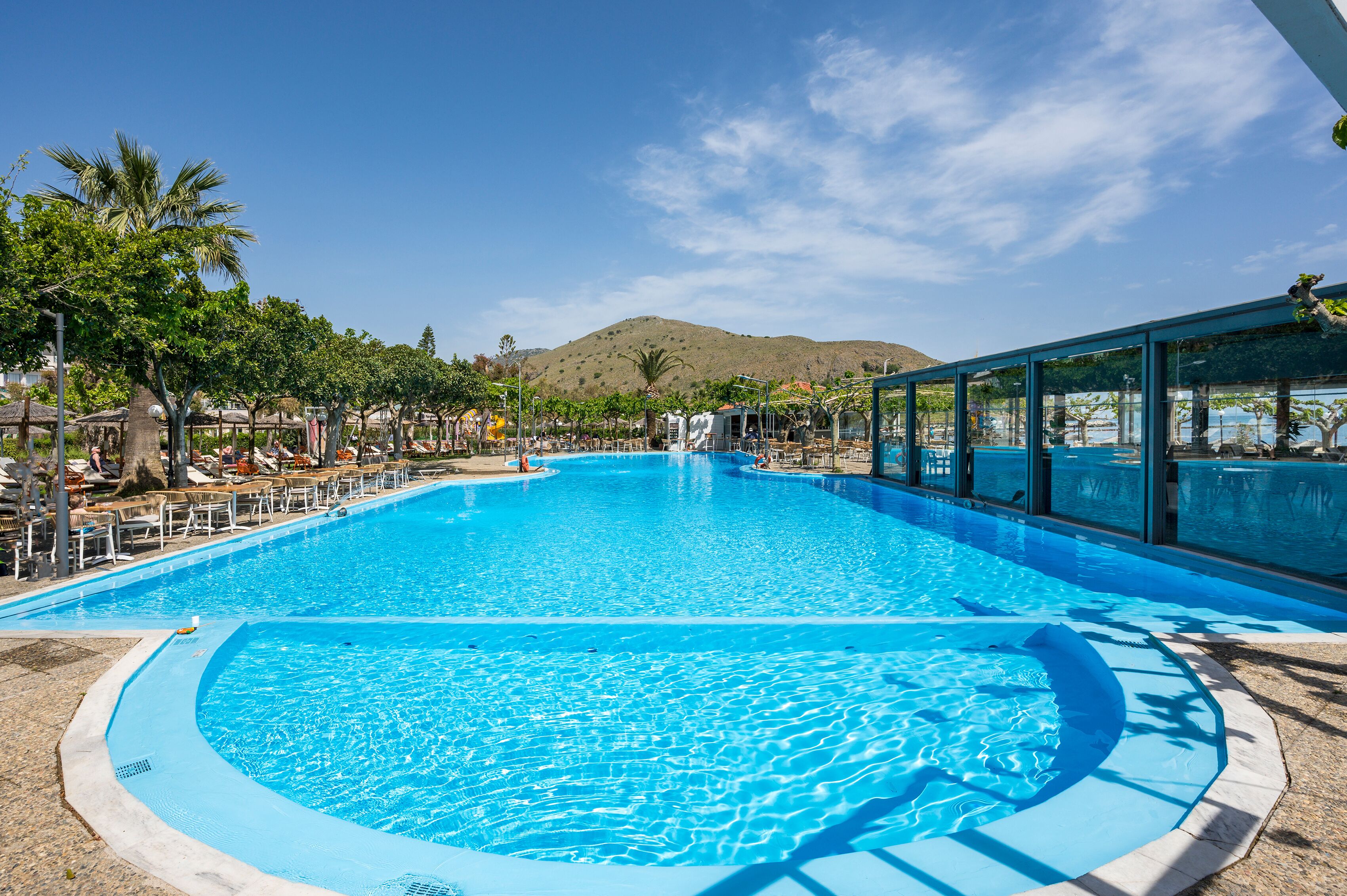 Une grande piscine d’un bleu éclatant, de forme circulaire, entourée de chaises longues et de palmiers, avec une montagne en arrière-plan.