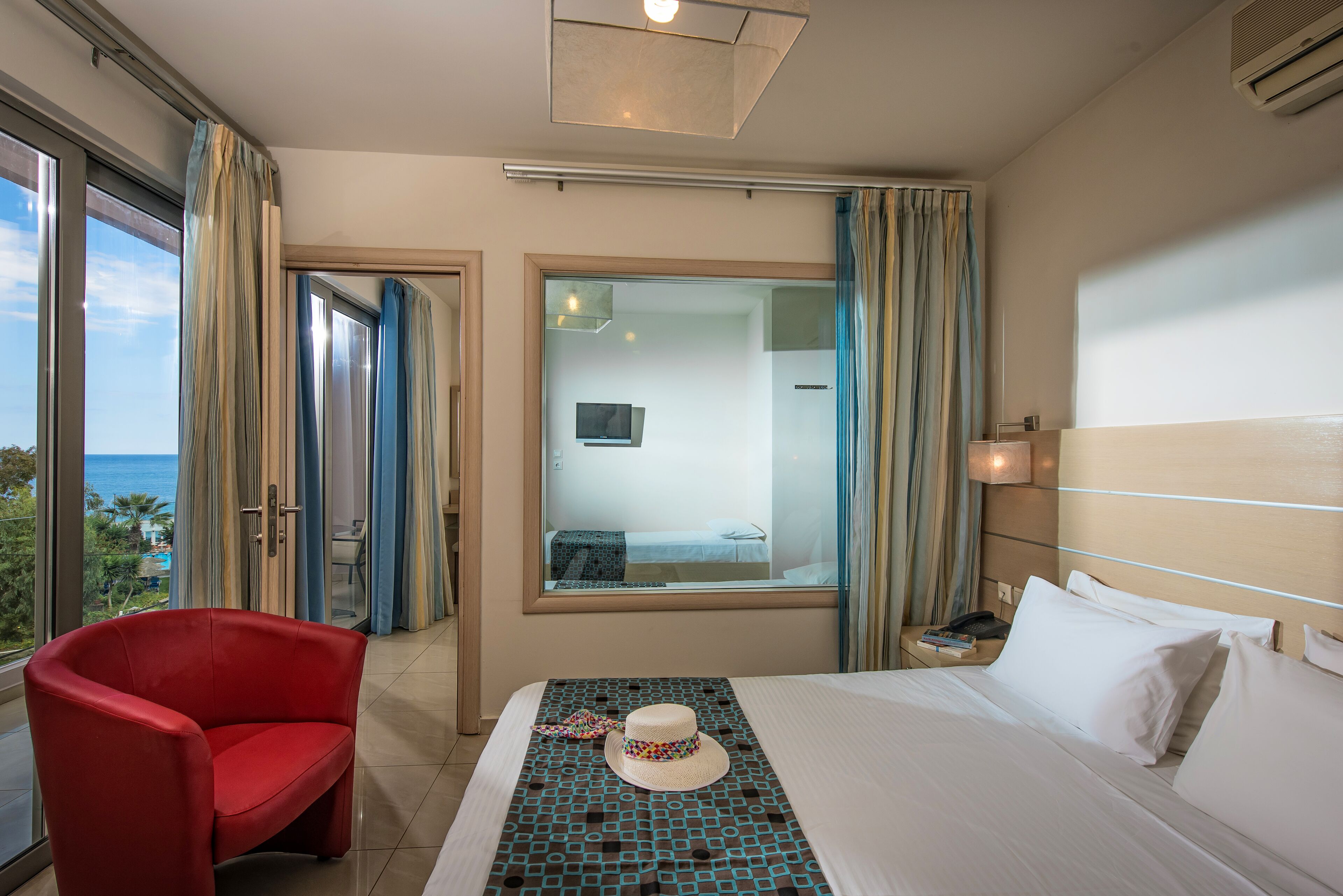 A hotel room with a bed, a red chair, a window with curtains, and a view of the ocean.