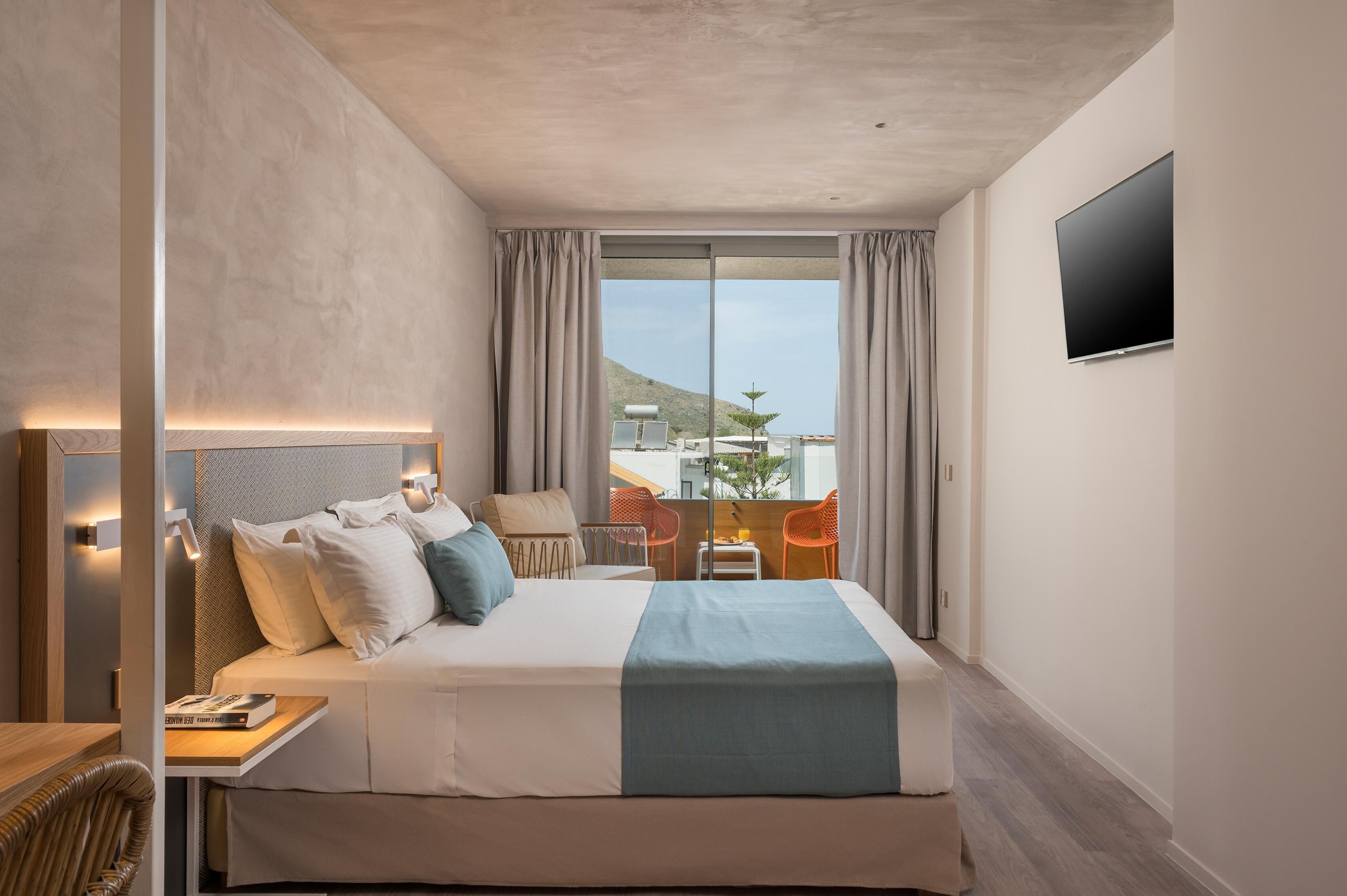 A hotel room with a large bed, a TV mounted on the wall, a balcony with chairs, and a view of the outside.