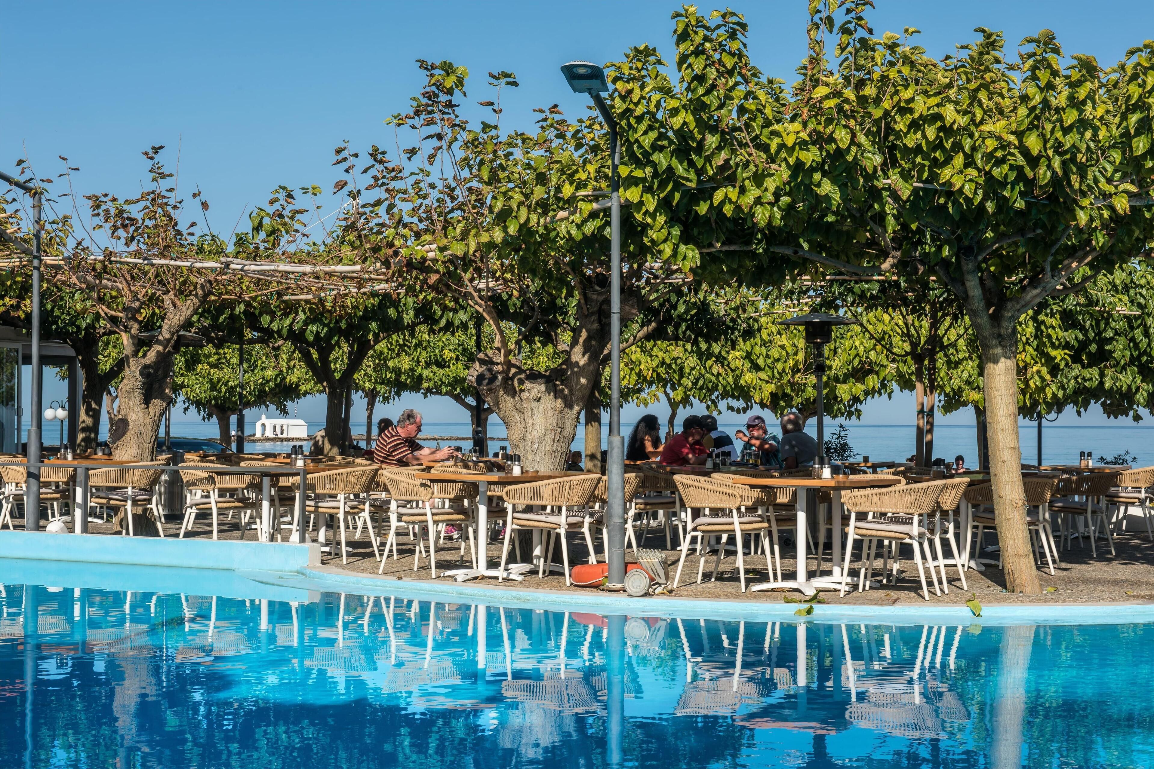 A poolside area with tables and chairs under trees, overlooking a body of water.