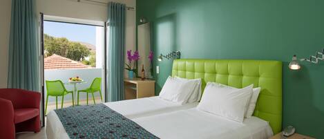 Habitación de hotel con un cabecero tapizado en verde, una cama grande con ropa blanca, un sillón rojo, una pequeña mesa redonda con sillas, y un balcón con vistas a árboles y un edificio.