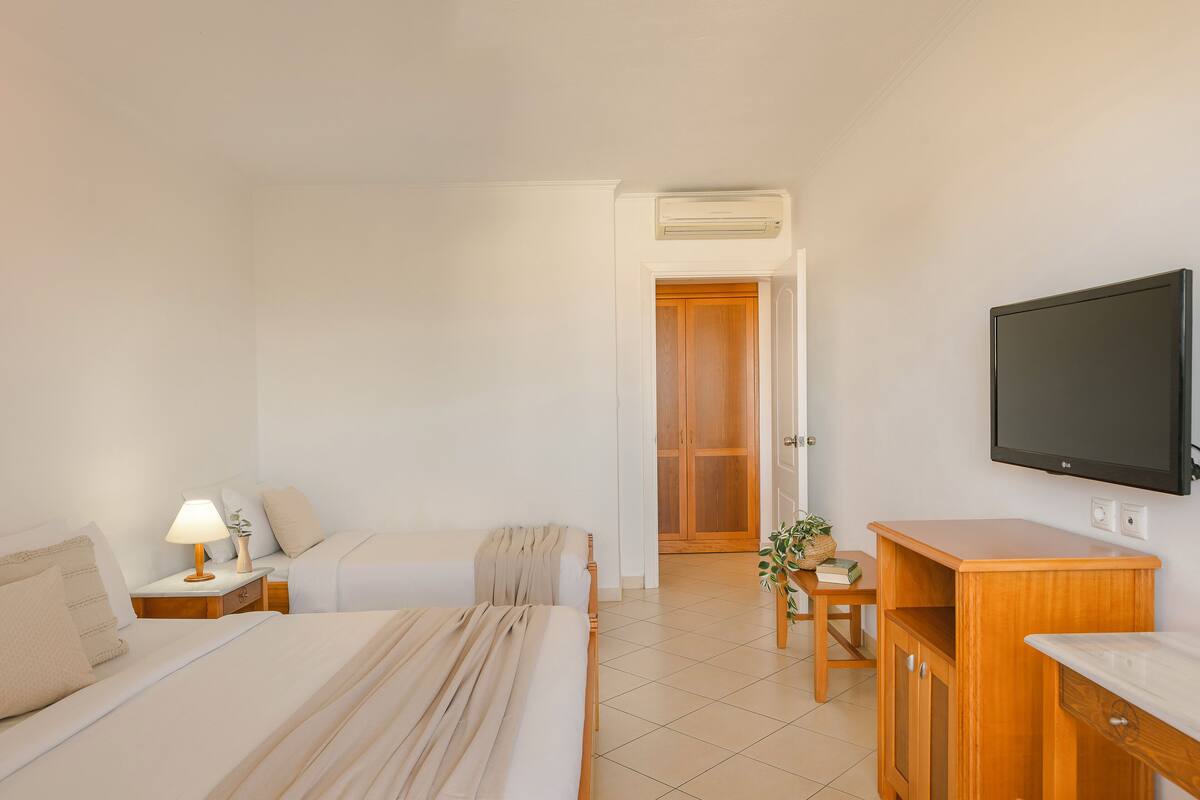 Una habitación de hotel con cama, mesita de noche con lámpara, televisor montado en la pared, aparador de madera y puerta que da a otra habitación.