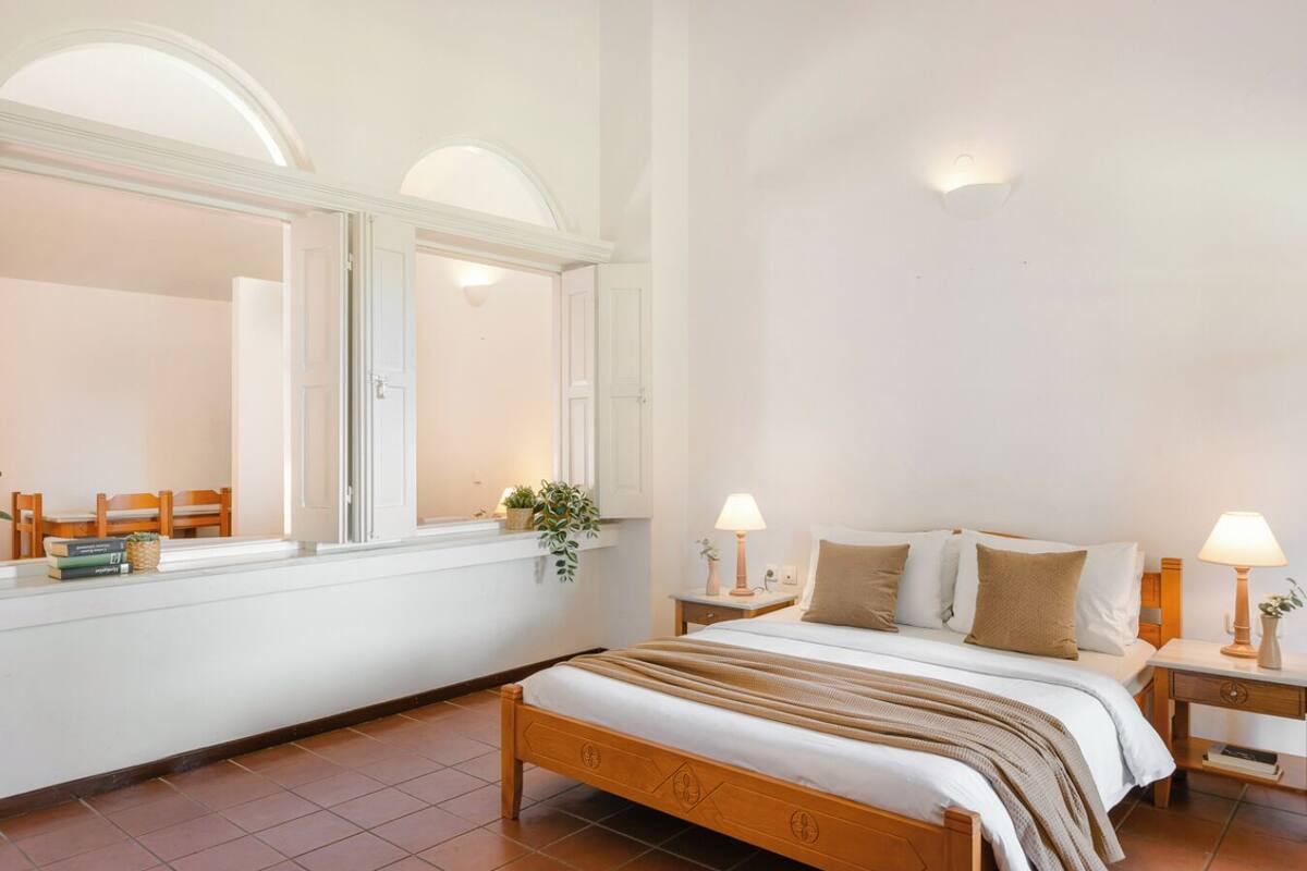 Un dormitorio con una cama de madera, dos mesitas de noche con lámparas, una ventana con vistas y una planta pequeña en el alféizar.