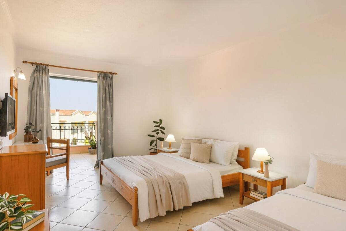 Habitación de hotel con cama, mesitas de noche, escritorio, silla, balcón con cortinas y vistas a edificios.