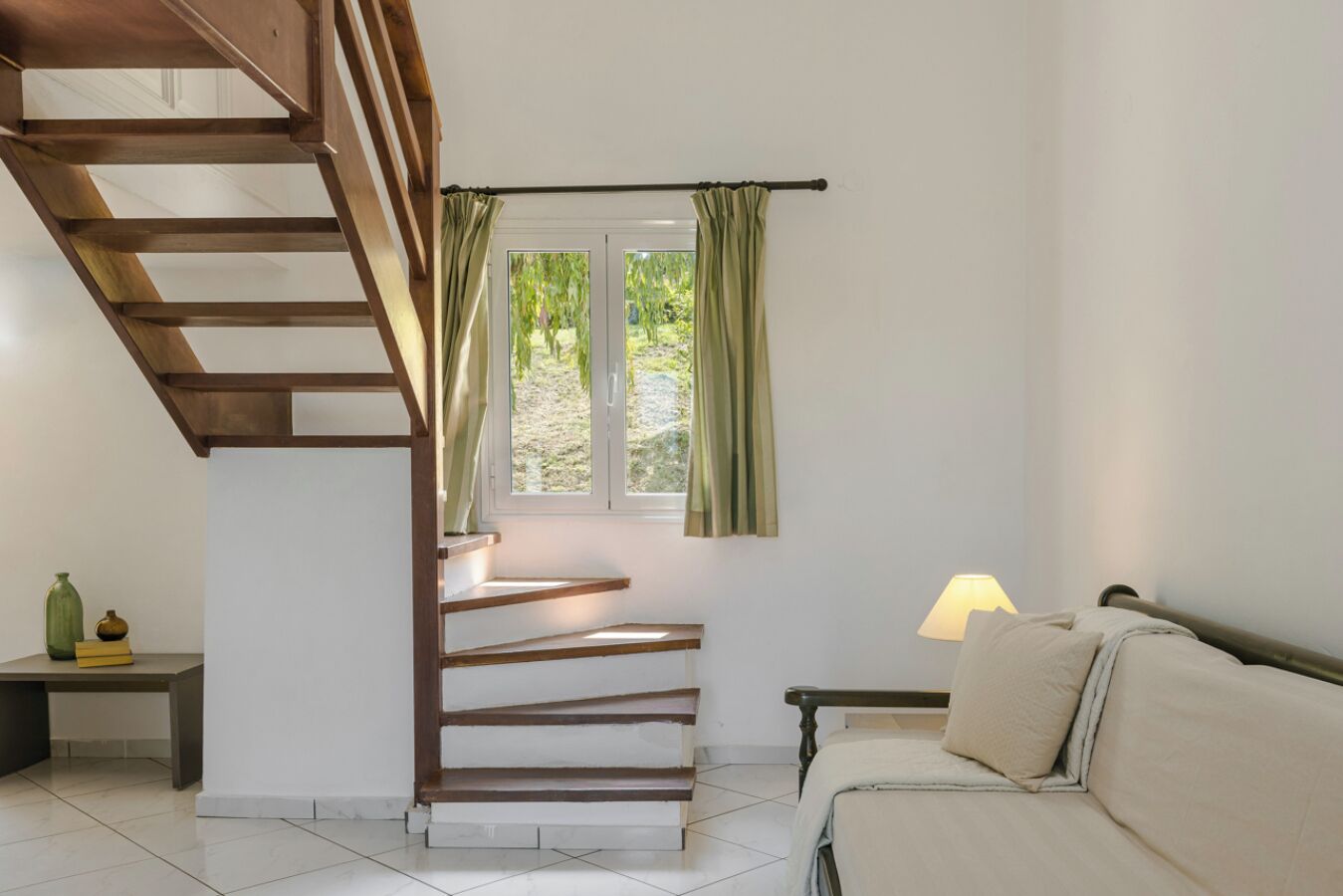 A staircase with wooden steps leading to a window with green curtains. A bed with a white pillow and a lamp on a bedside table are visible.