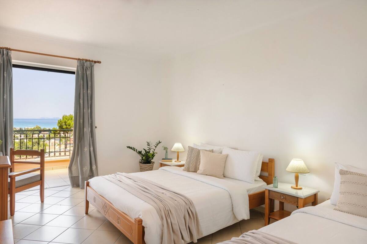 Un dormitorio con una cama grande, dos mesitas de noche con lámparas, una silla y vistas al mar desde un balcón.
