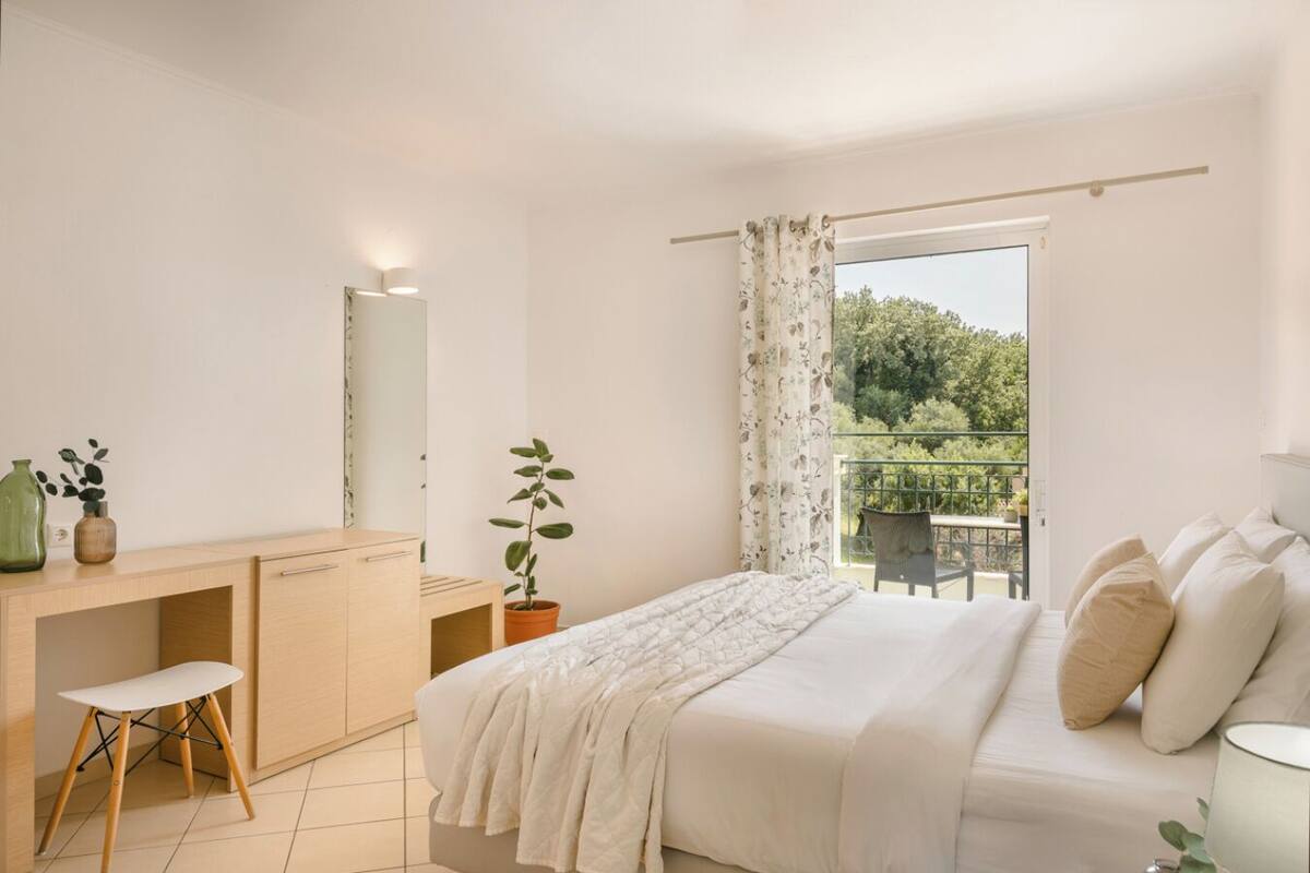 Un dormitorio con cama, escritorio, silla, espejo y balcón con vistas a zonas verdes.