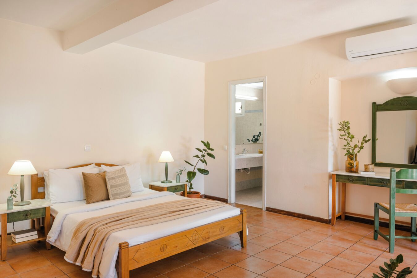 Une chambre à coucher comprenant un lit, des tables de chevet, un bureau, un miroir et une salle de bain visible à travers une porte ouverte.