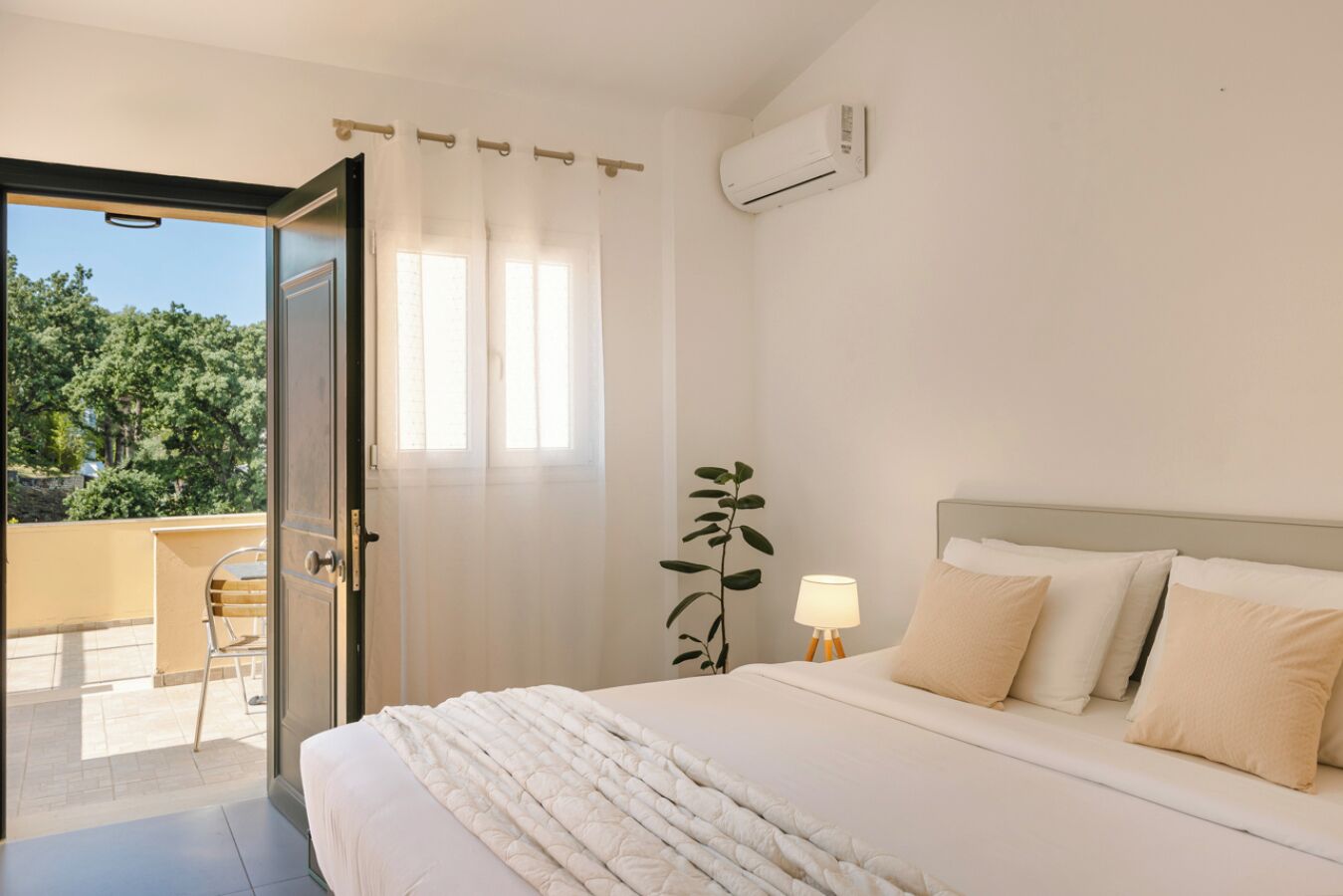 A hotel room with a bed, a bedside lamp, a potted plant, and a view of the outdoors.