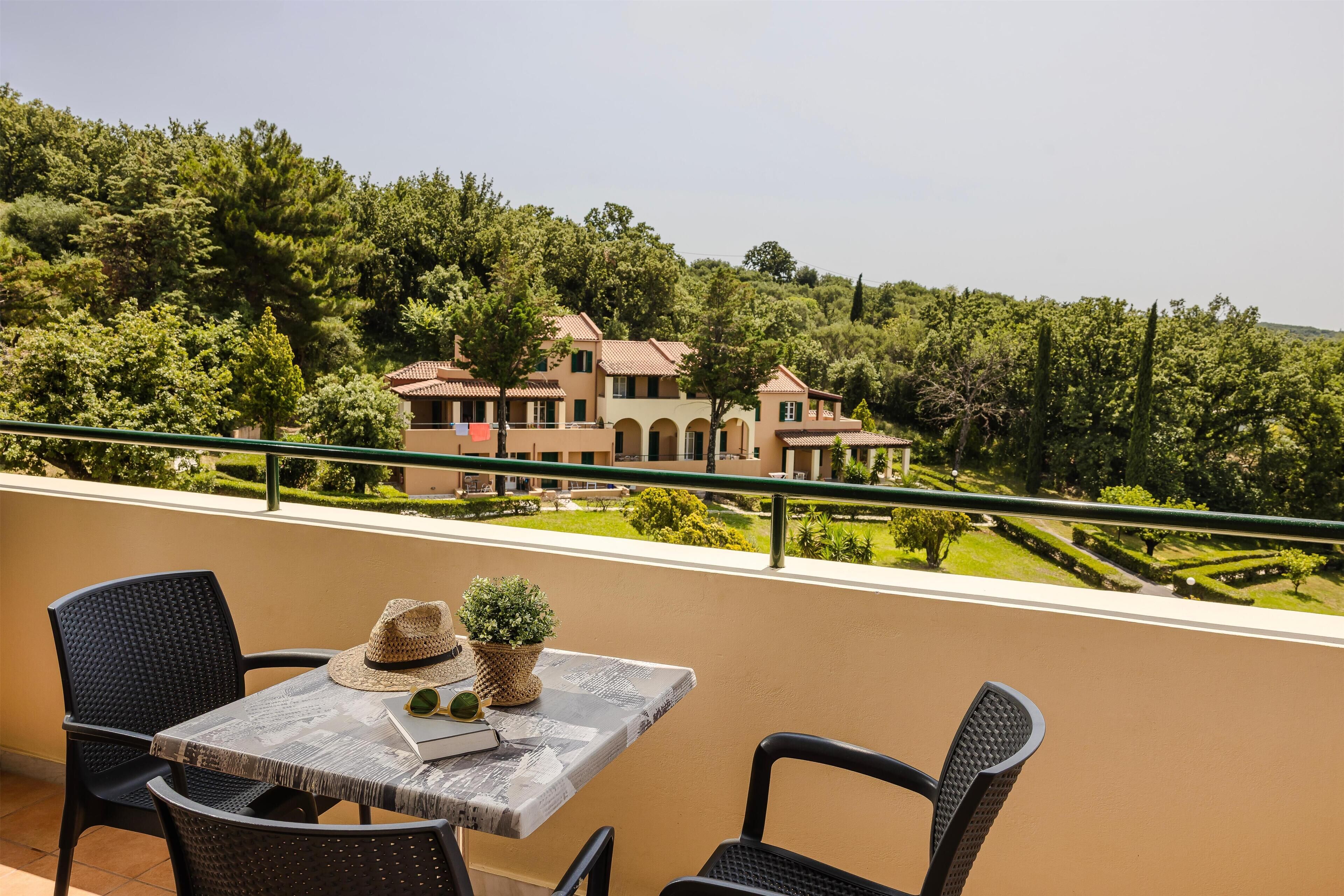 Ein Balkon mit Tisch und Stühlen, der auf eine üppige, grüne Landschaft und ein großes Gebäude mit Ziegeldach blickt.