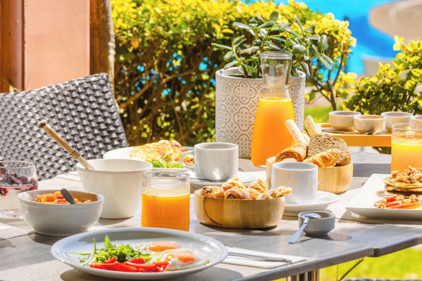 A table set with breakfast items including eggs, toast, fruit, and juice, outdoors with a view of greenery and a pool.