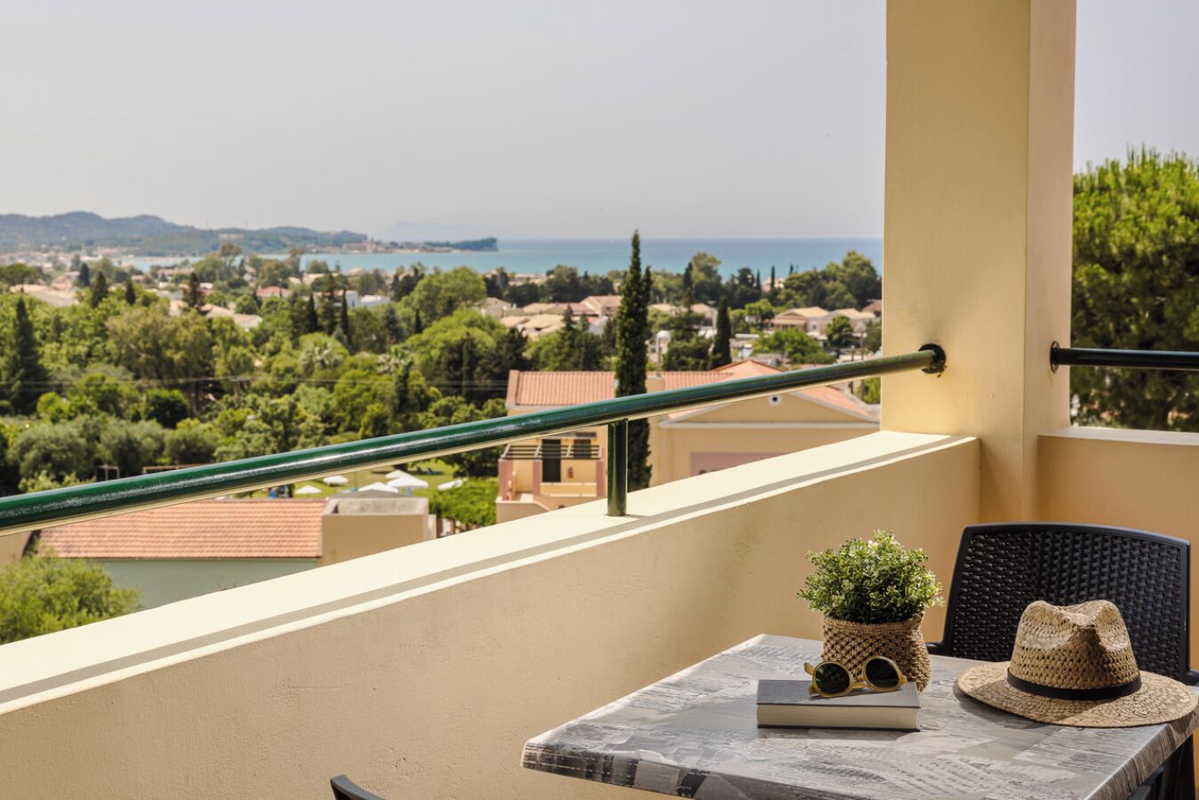 A balcony with a view of a coastal town, a table with a book, a potted plant, and a hat.