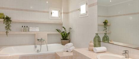 A modern bathroom with a bathtub, a sink, and a large mirror. There are plants and toiletries on the shelves.