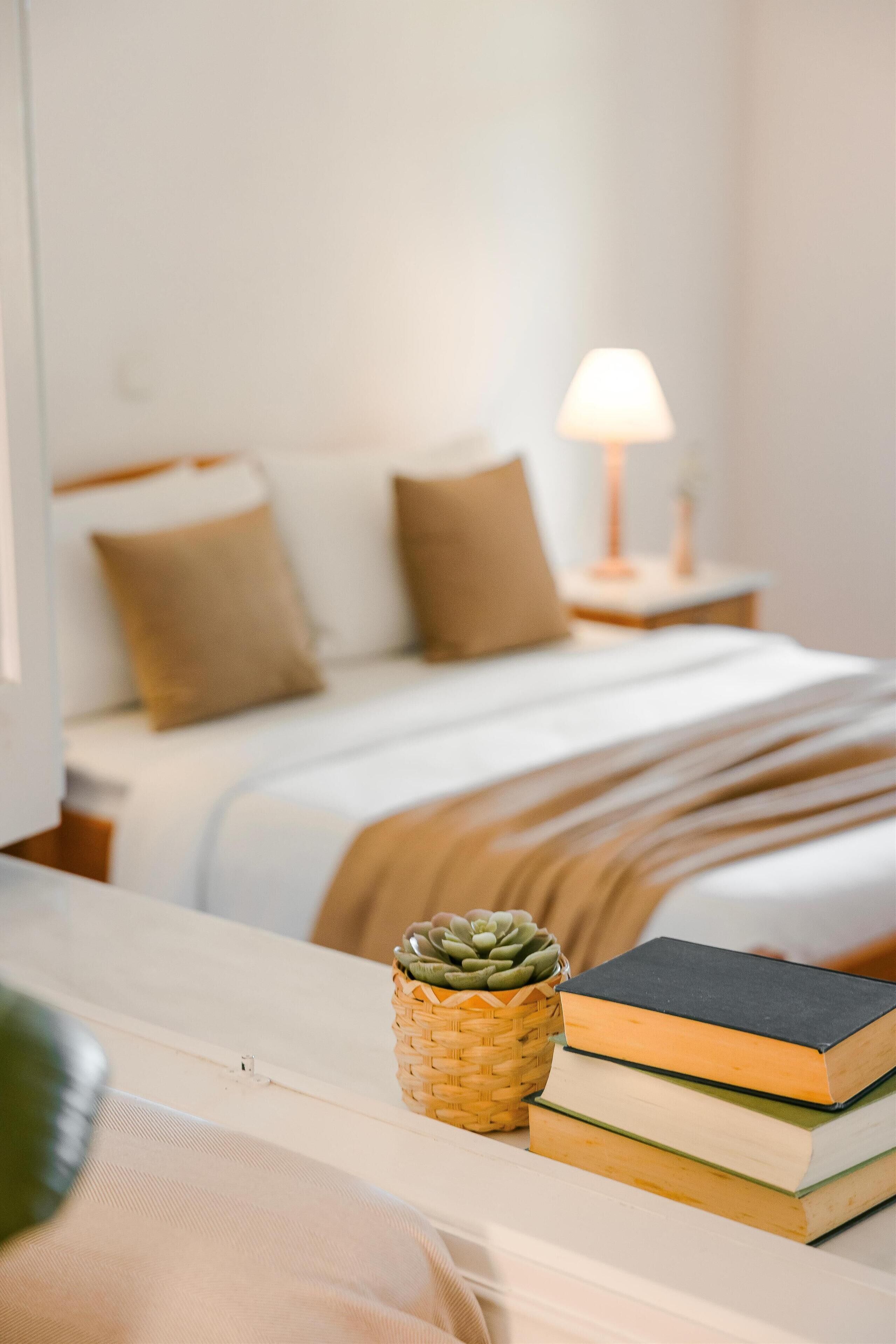 Una cama bien hecha con edredón blanco y dos almohadas, una lámpara de noche, una pequeña cesta tejida con una suculenta, y una pila de libros en una mesita de noche.