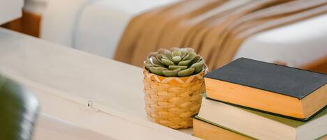 A neatly made bed with a white comforter and two pillows, a bedside lamp, a small woven basket with a succulent plant, and a stack of books on a bedside table.