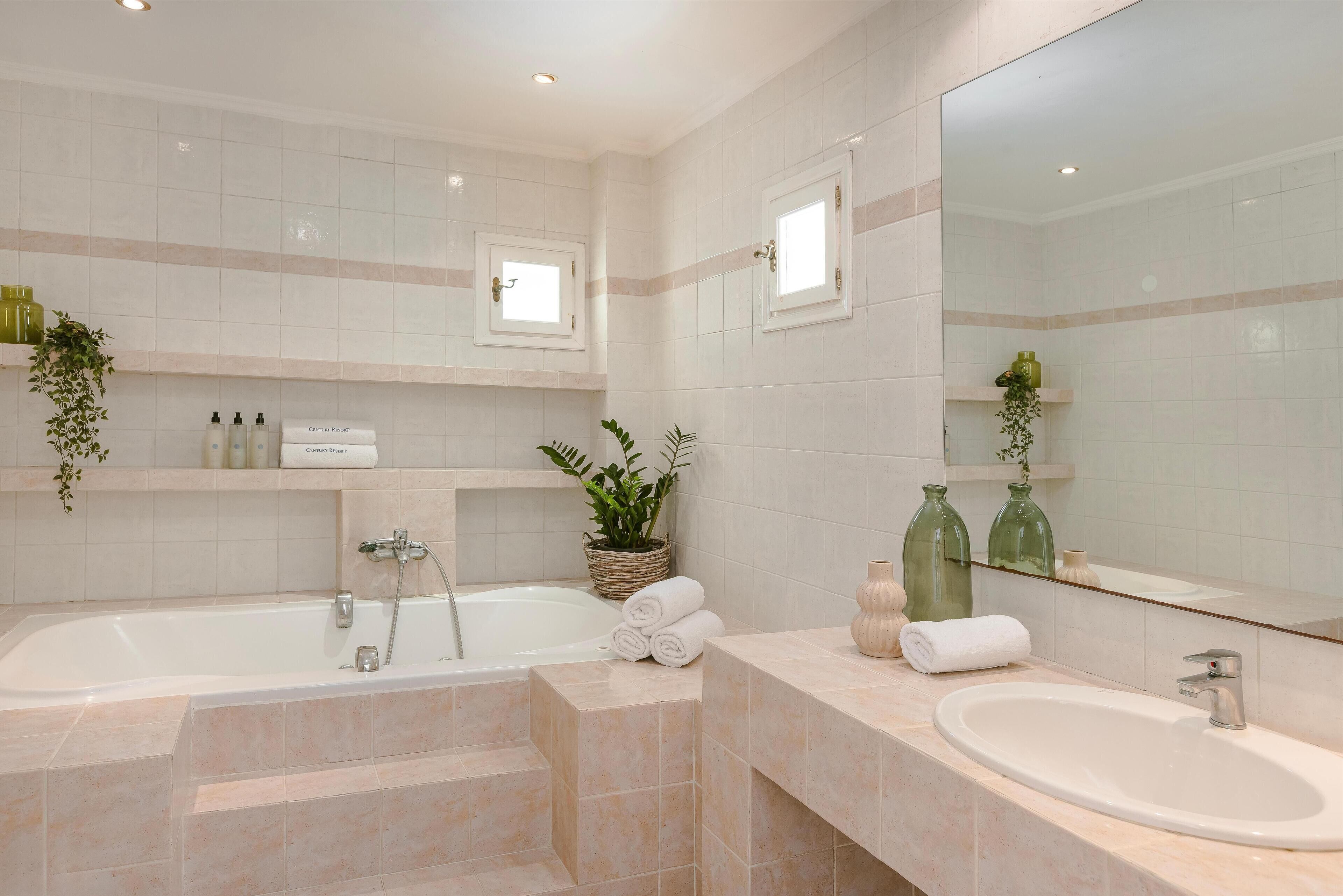 Un baño moderno con bañera, lavamanos y un espejo grande. Hay repisas con artículos de aseo personal y una planta en maceta.