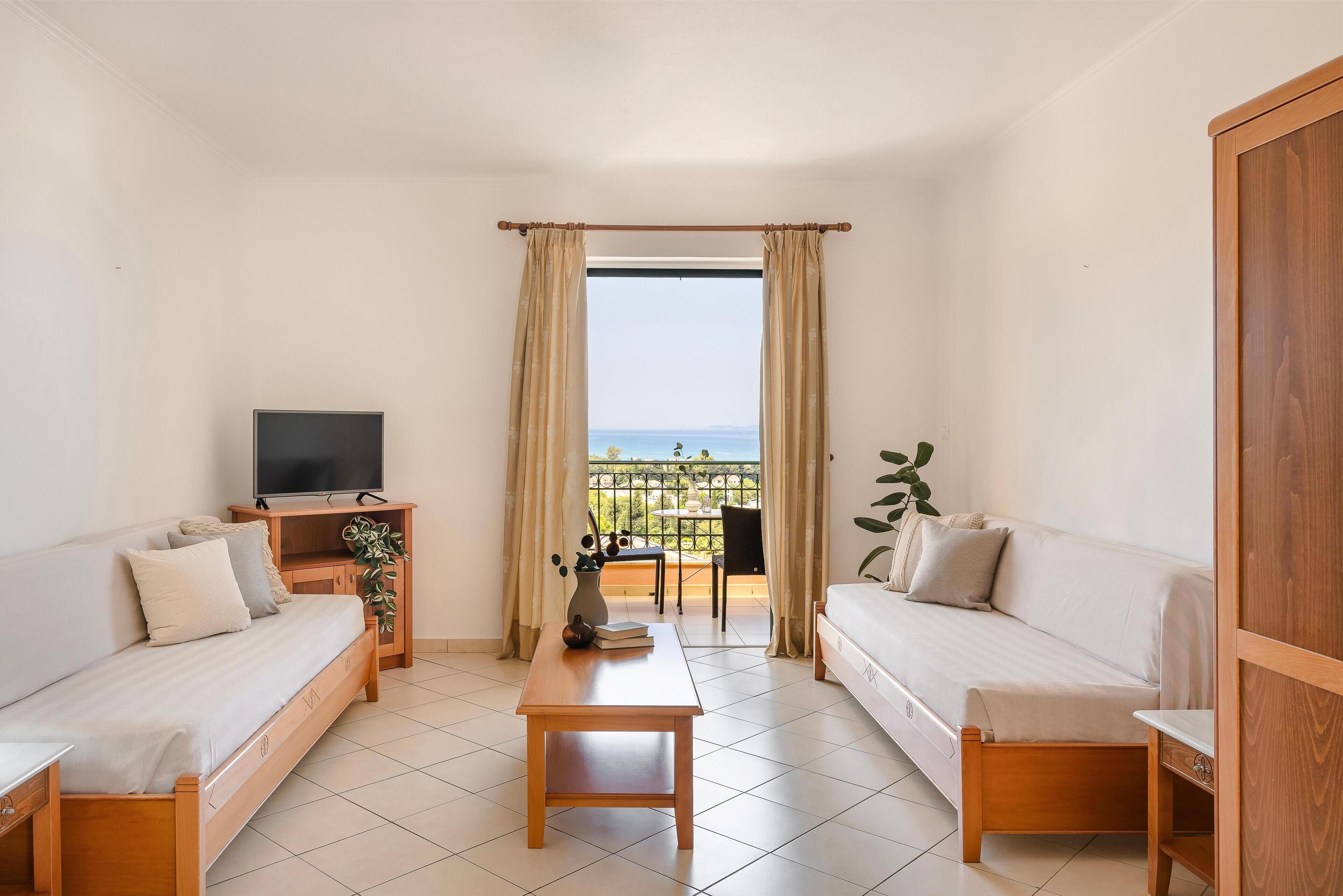 A modern, well-lit room with a sofa, a small table, and a TV on a wooden stand. A balcony with a view of the sea is visible through a window with curtains.