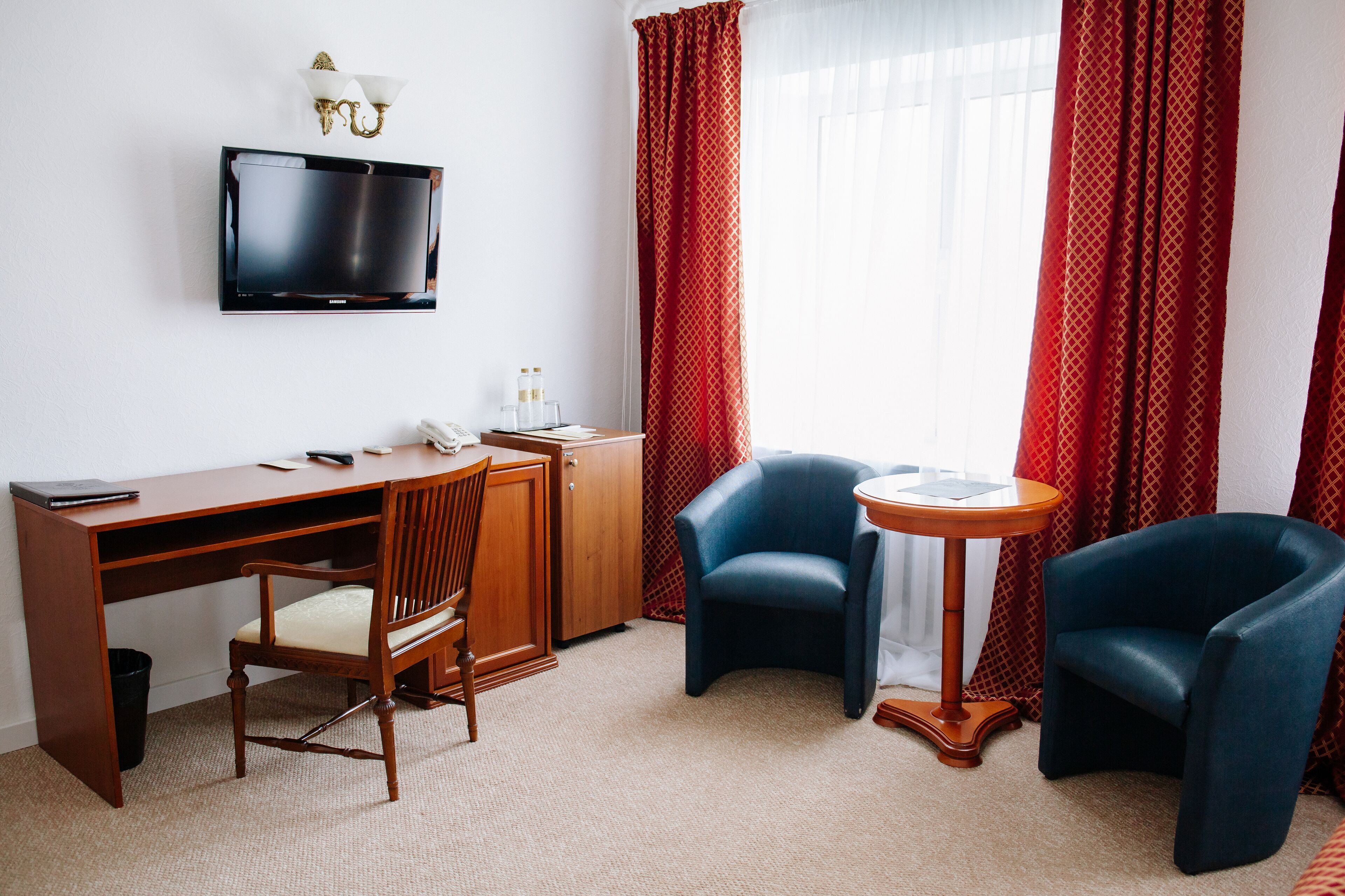 A hotel room with a wooden desk, two blue armchairs, a small round table, a TV mounted on the wall, and red patterned curtains.