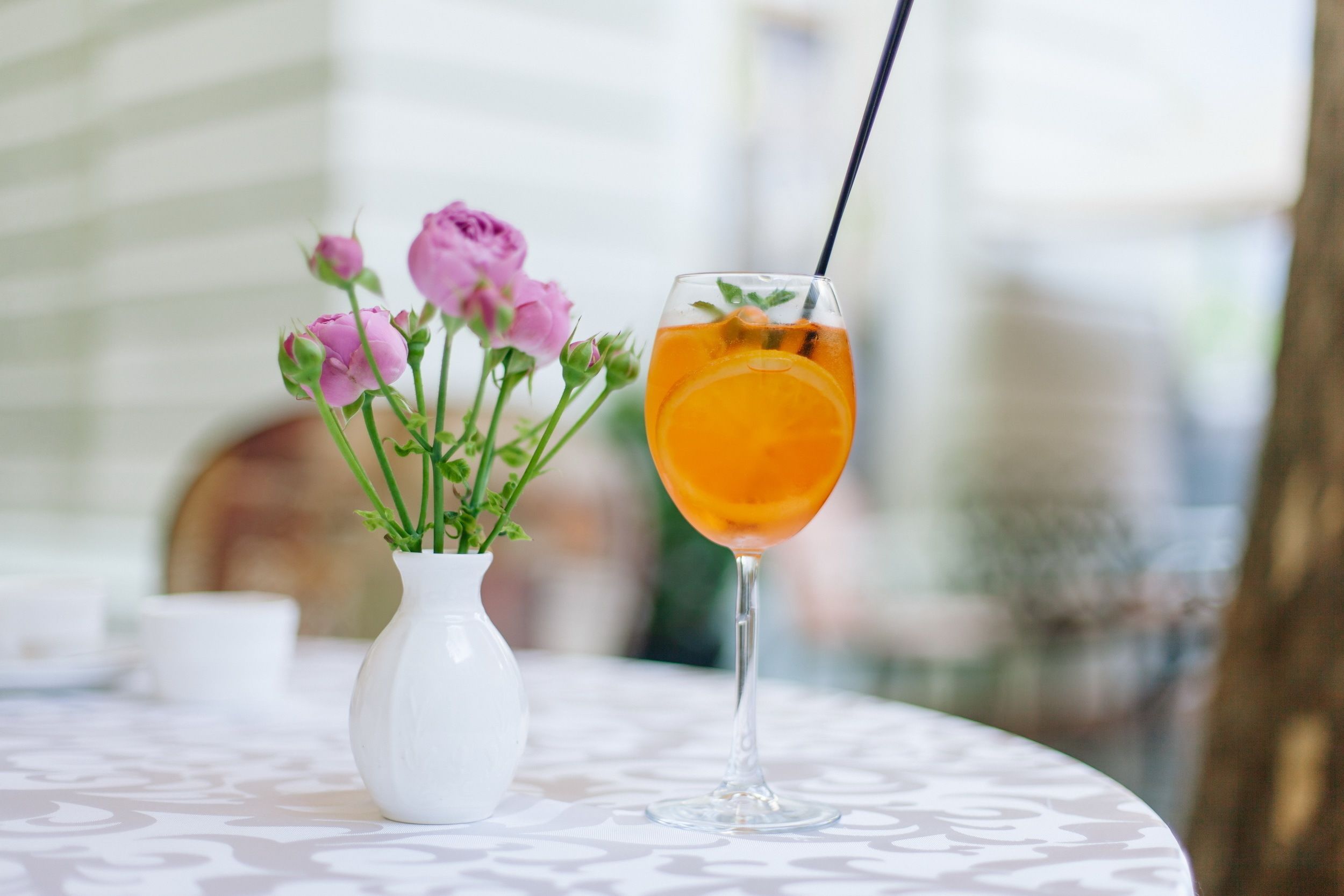 Una mesa con un jarrón blanco lleno de flores rosas y un vaso de bebida naranja.