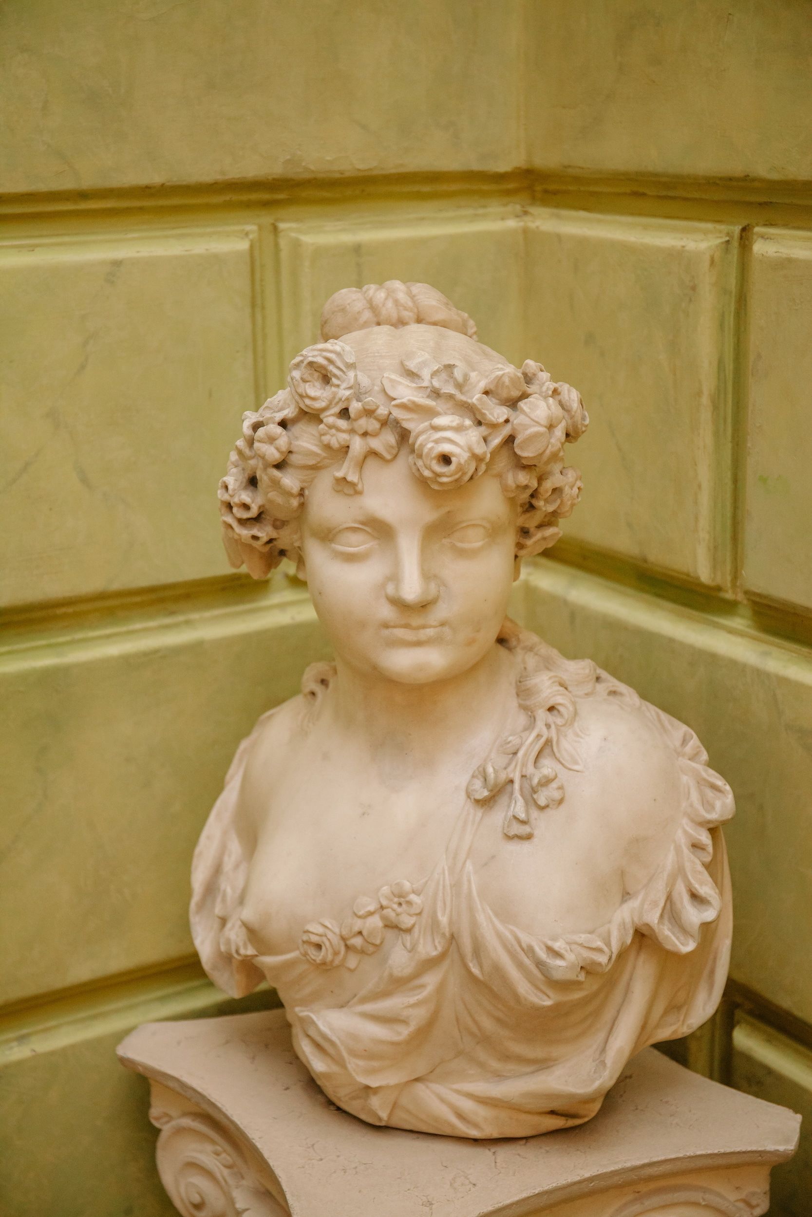 Una escultura de busto con adornos florales sobre un pedestal frente a un muro con paneles.
