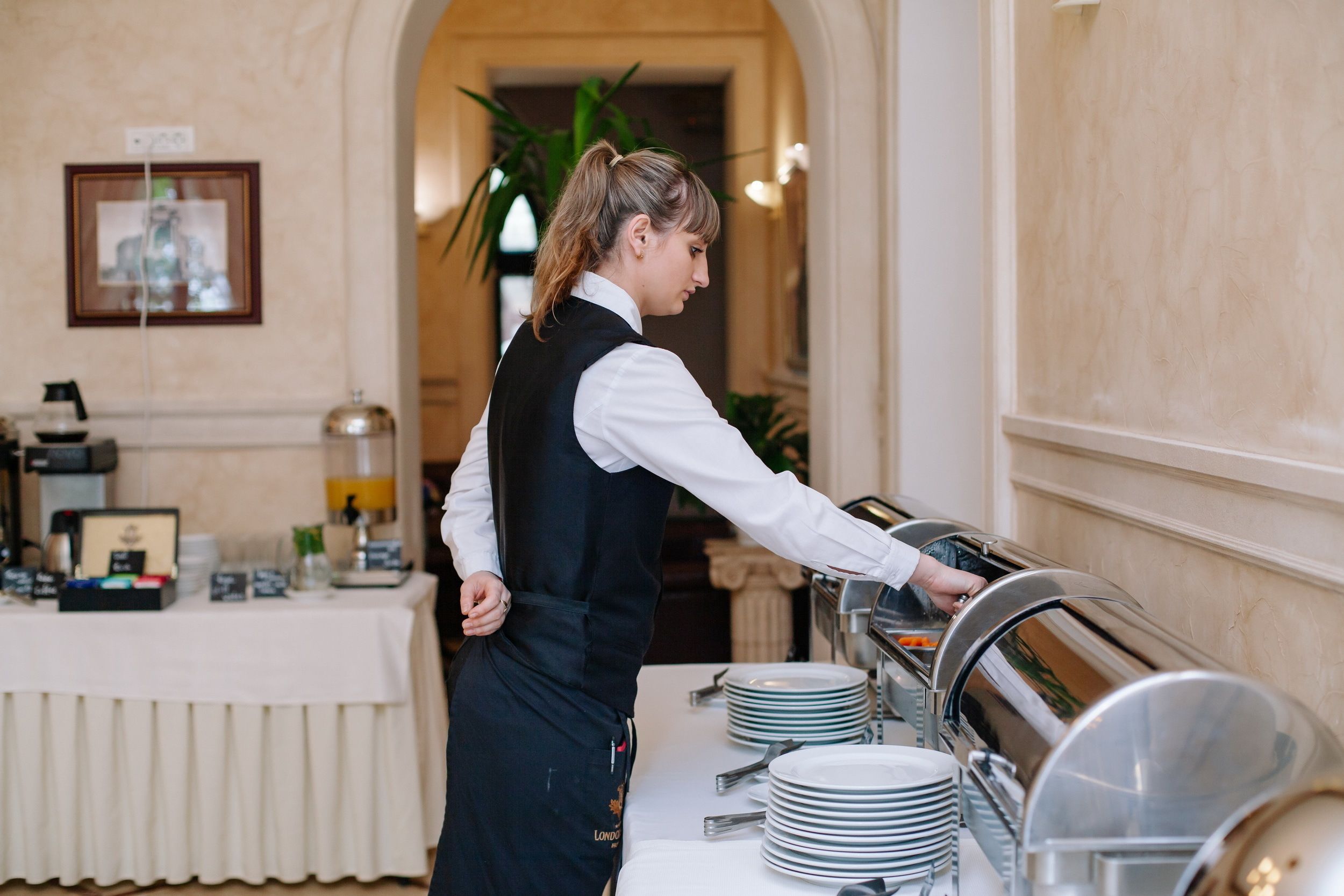 A buffet setup with a server serving food, a white tablecloth, and a buffet line with plates and silverware.