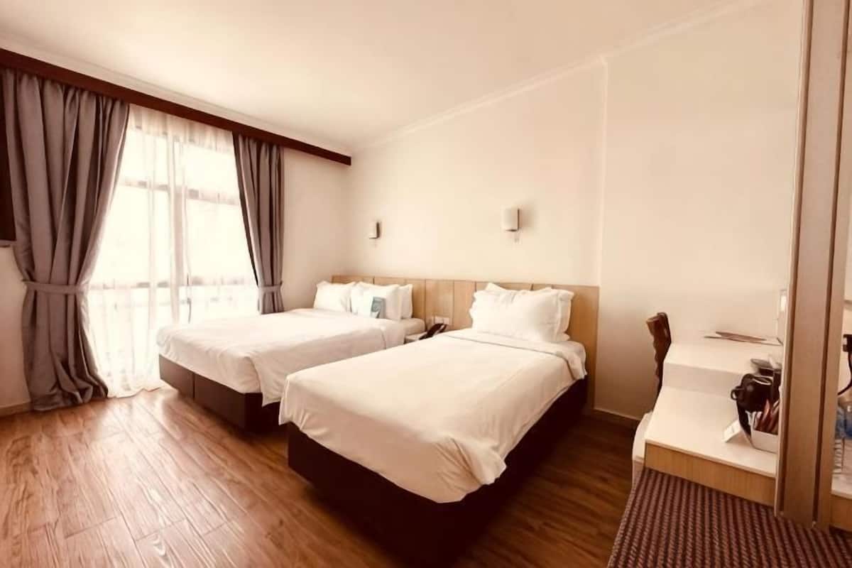 Een hotelkamer met twee bedden, een houten vloer, een bureau met stoel en een raam met gordijnen.