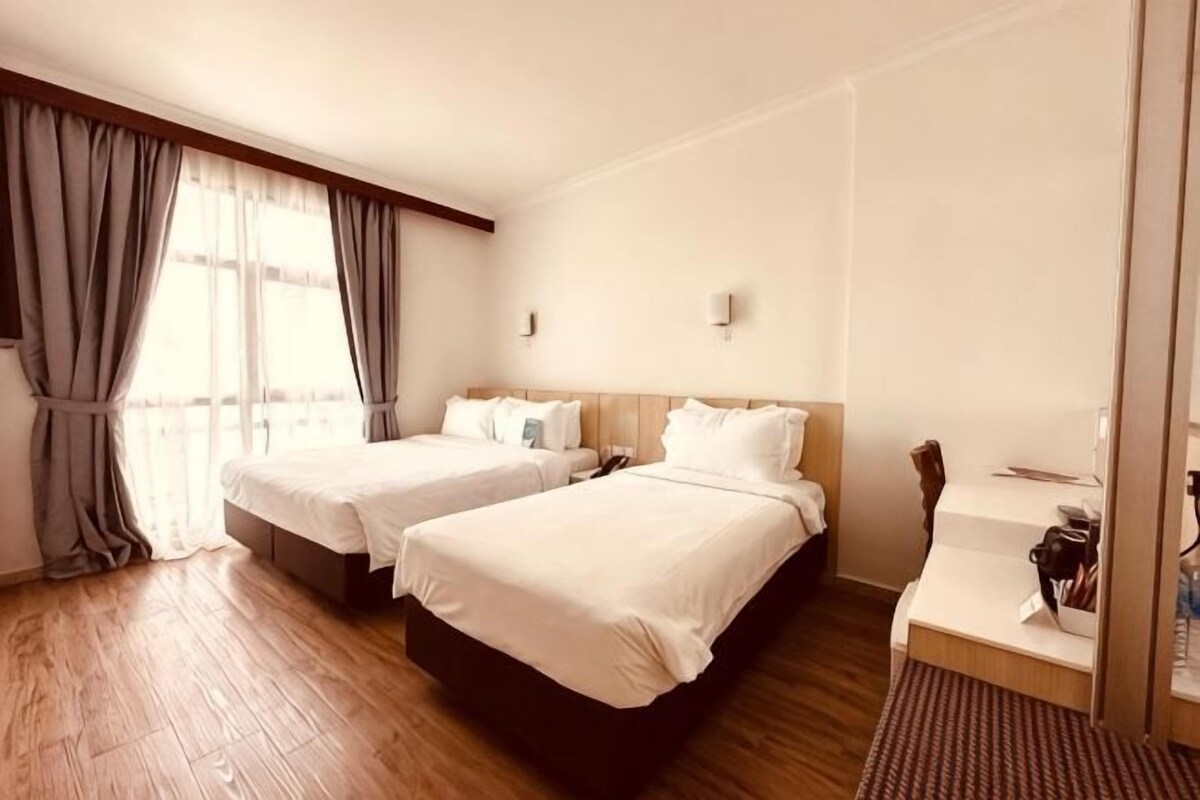 A hotel room with two beds, a wooden floor, a desk with a chair, and a window with curtains.