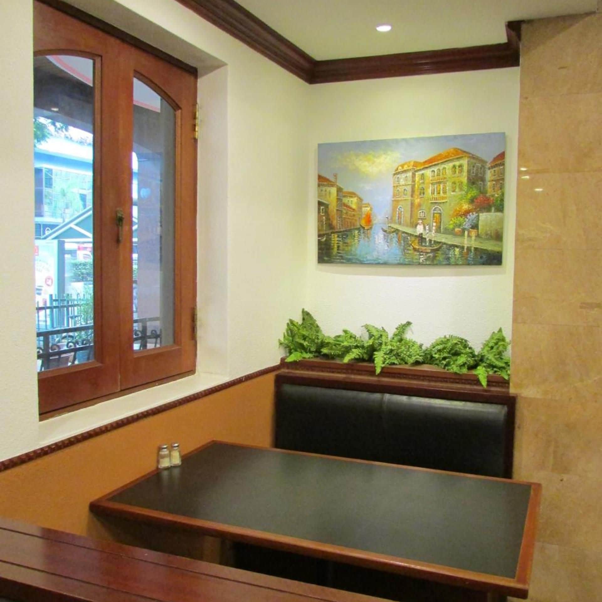 Un área de comedor con un banco y una mesa, una ventana con vistas a edificios, y un cuadro de una escena de canal en la pared.