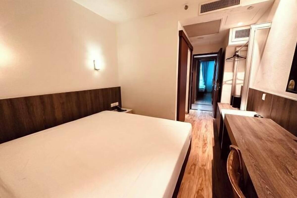 Een hotelkamer met een bed, een houten bureau, een spiegel en een gang die naar een andere kamer leidt.