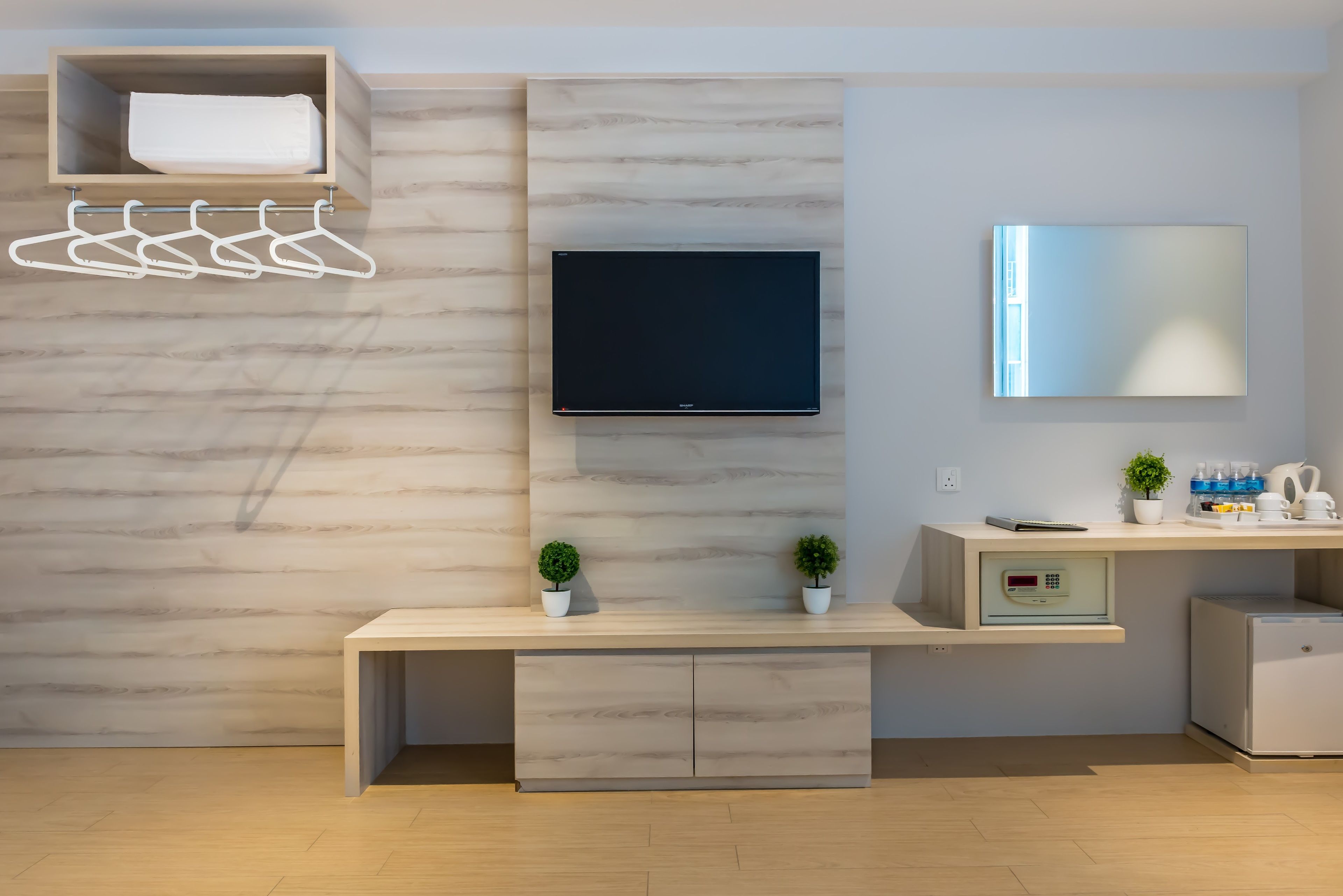 A modern hotel room with a wooden wardrobe, a flat-screen TV, a mirror, a small desk with a plant, and a mini-fridge.