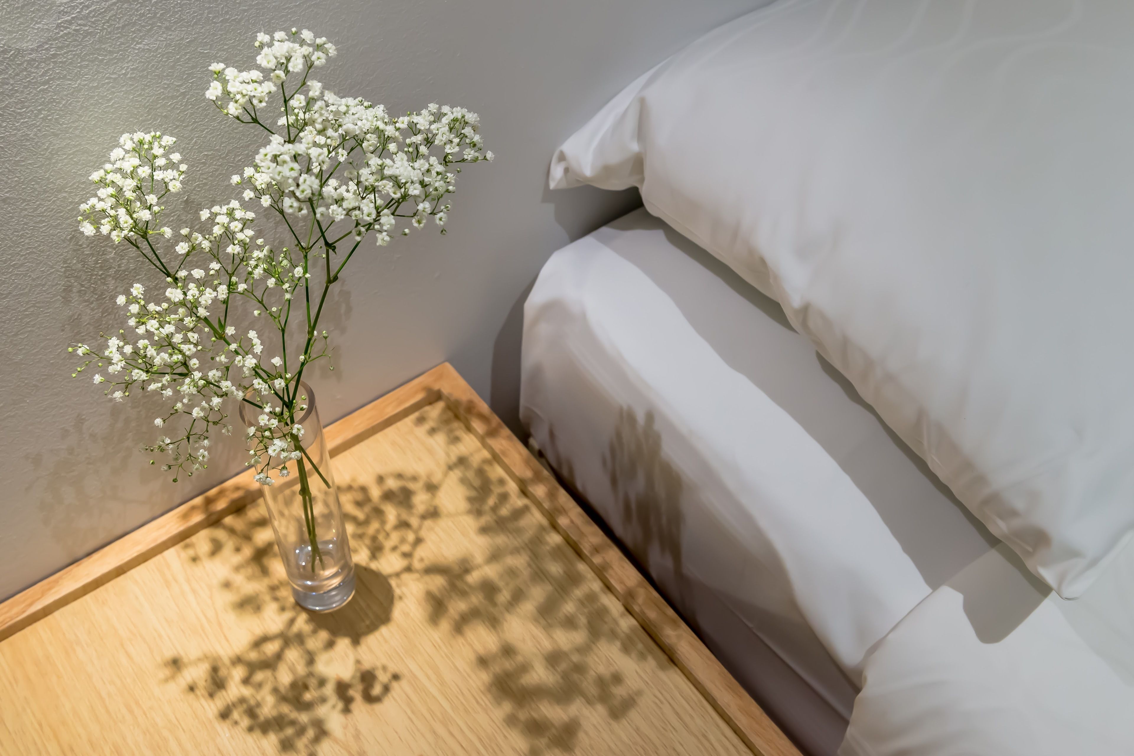 A wooden nightstand with a clear vase containing white flowers, positioned next to a bed with white bedding.