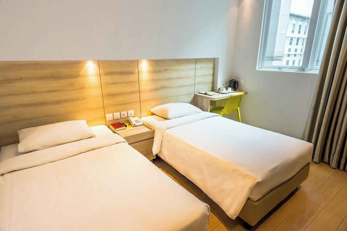 A hotel room with two beds, a desk with a chair, a window with curtains, and a view of buildings outside.