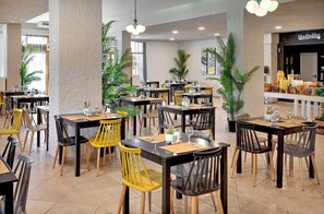 A restaurant with multiple tables and chairs, some with yellow chairs, and a bar area in the background. - The Grove Seaside Hotel (Nafplio)