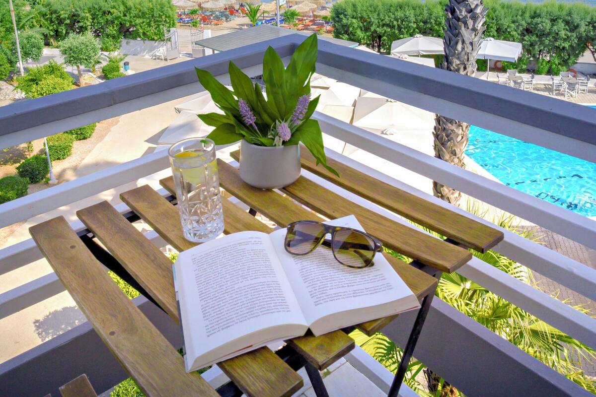 A wooden table with a book, a glass of water, and a potted plant, overlooking a pool and a beach with clear blue water.