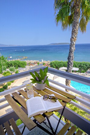 Ett träbord med en bok, ett glas vatten och en krukväxt, med utsikt över en pool och en strand med kristallklart blått vatten.