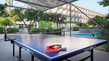 An outdoor ping pong table with a net, surrounded by greenery and a building in the background.