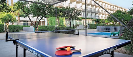 Una mesa de ping pong al aire libre con red, rodeada de follaje y un edificio en el fondo.