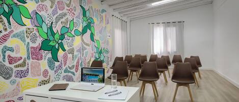 Une salle de conférence avec un bureau blanc, un ordinateur portable et un verre d’eau. Les murs de la salle sont décorés de motifs et les chaises sont marron.