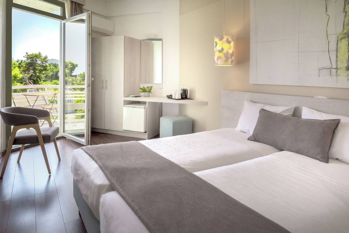 A modern hotel room with a large bed, a desk with a chair, and a balcony with a view of trees and mountains.