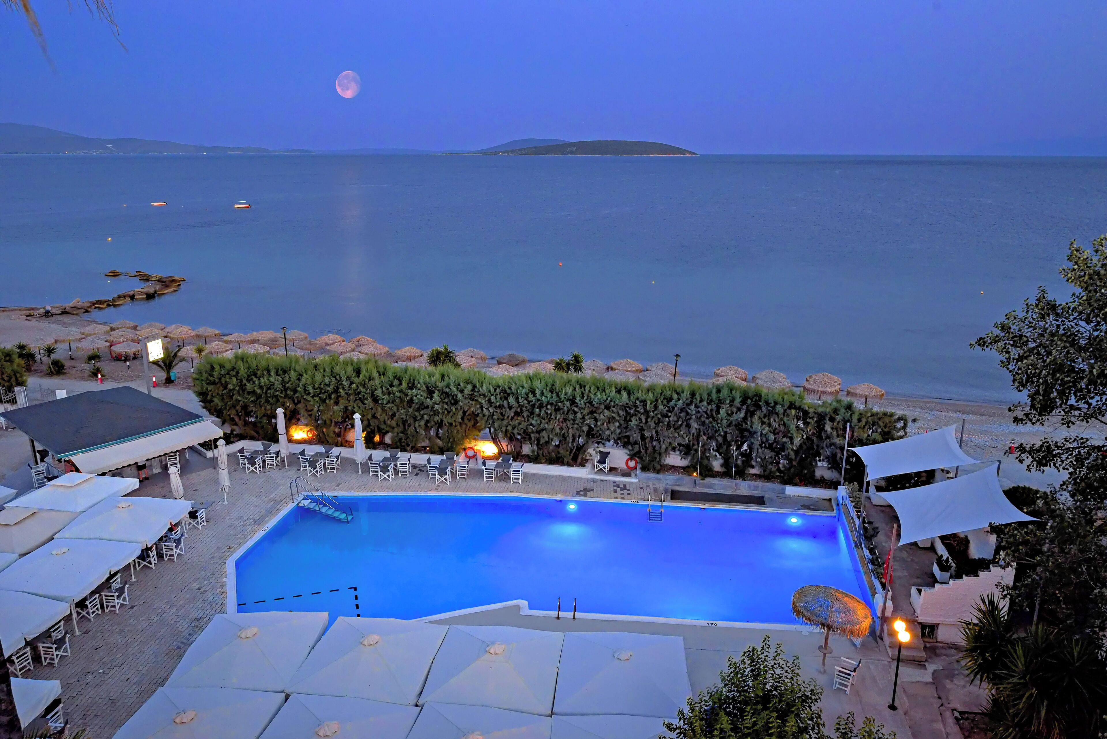 A pool area with lounge chairs and umbrellas, overlooking a body of water with a moon in the sky.