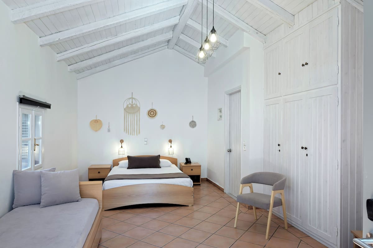 A spacious bedroom with a large bed, a chair, a wardrobe, and a ceiling with exposed beams.